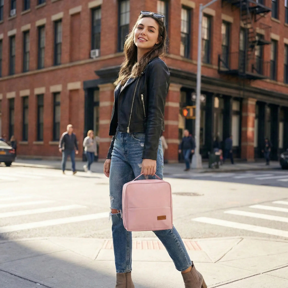A smiling young woman in a black leather jacket and ripped jeans walking on a city street, carrying a pastel pink Sunday Status stand-up travel makeup bag. The image highlights the bag as a trendy fashion accessory for modern urban life.