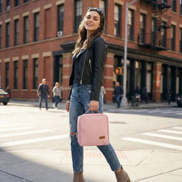 A smiling young woman in a black leather jacket and ripped jeans walking on a city street, carrying a pastel pink Sunday Status stand-up travel makeup bag. The image highlights the bag as a trendy fashion accessory for modern urban life.