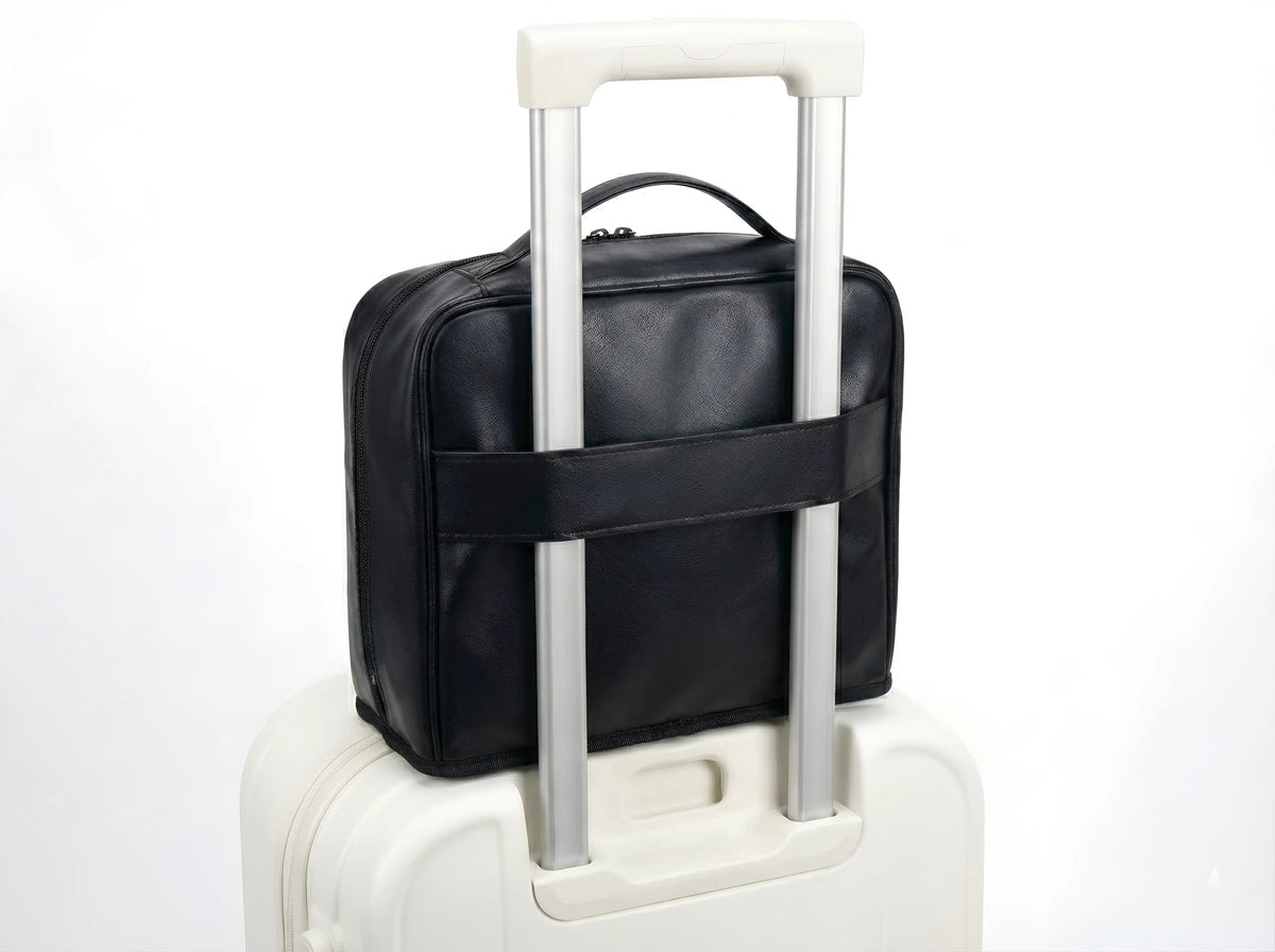 A close-up back view of the black Sunday Status travel makeup bag, showing the sturdy built-in luggage sleeve securely fitted over the handle of a white rolling suitcase for easy, hands-free travel