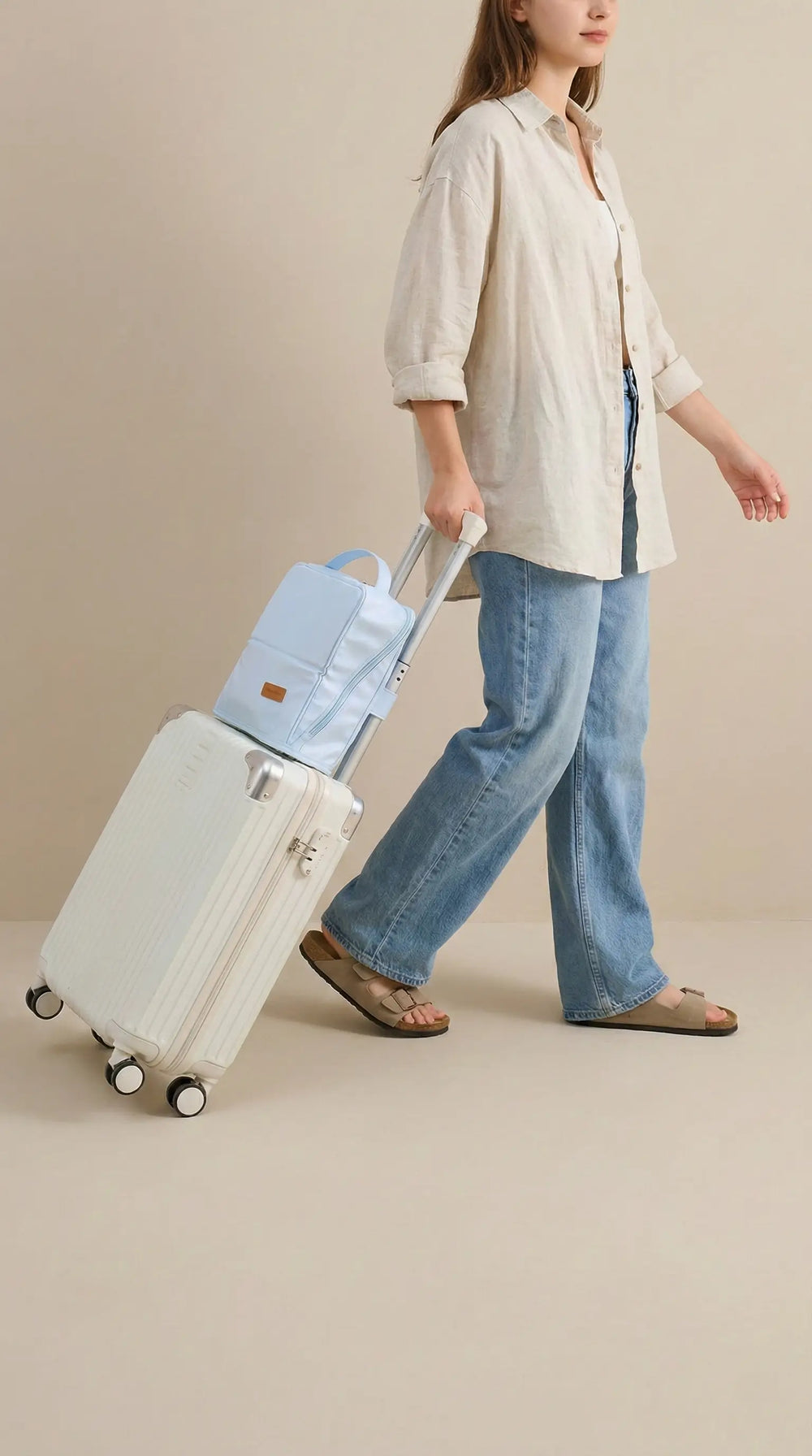 A woman in casual travel attire walking and pulling a white rolling carry-on suitcase. A light blue Sunday Status travel makeup bag is securely attached to the suitcase handle using its built-in luggage sleeve for hands-free travel