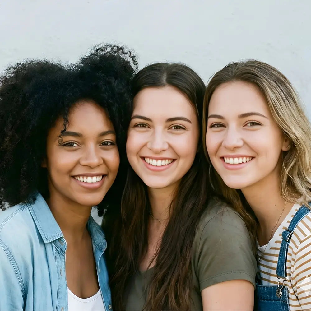 A close-up horizontal banner of three diverse young women smiling joyfully and leaning together against a neutral background, representing the inclusive and happy community of the Sunday Status brand