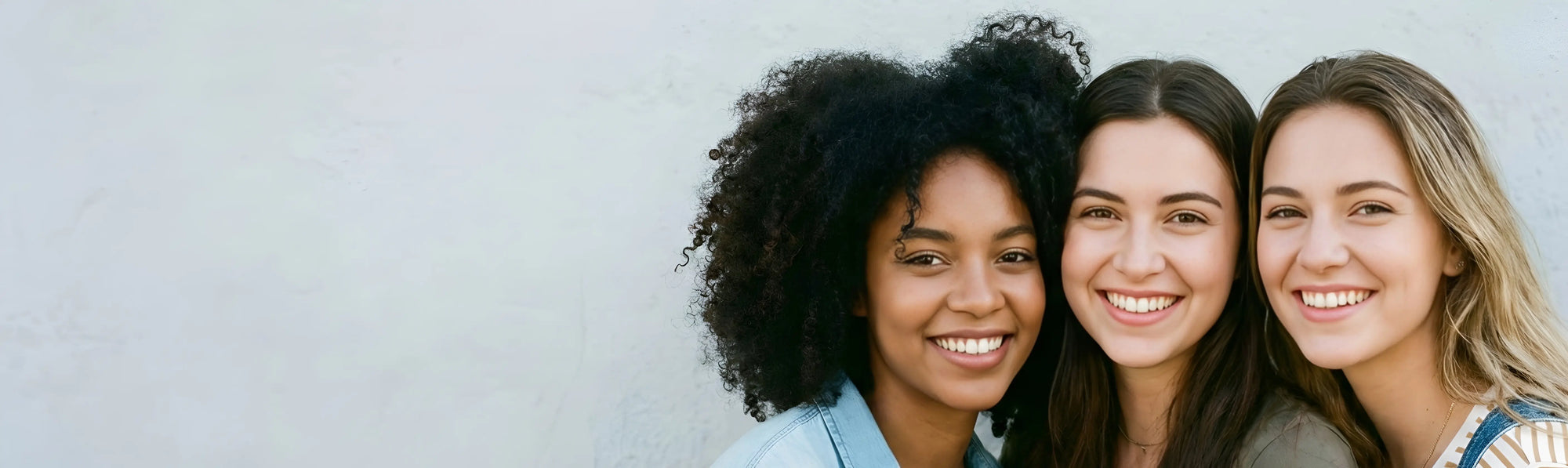 A close-up horizontal banner of three diverse young women smiling joyfully and leaning together against a neutral background, representing the inclusive and happy community of the Sunday Status brand